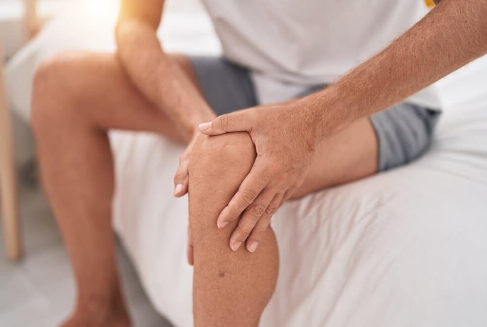 A person sitting on a bed, holding their knee, indicating pain or discomfort